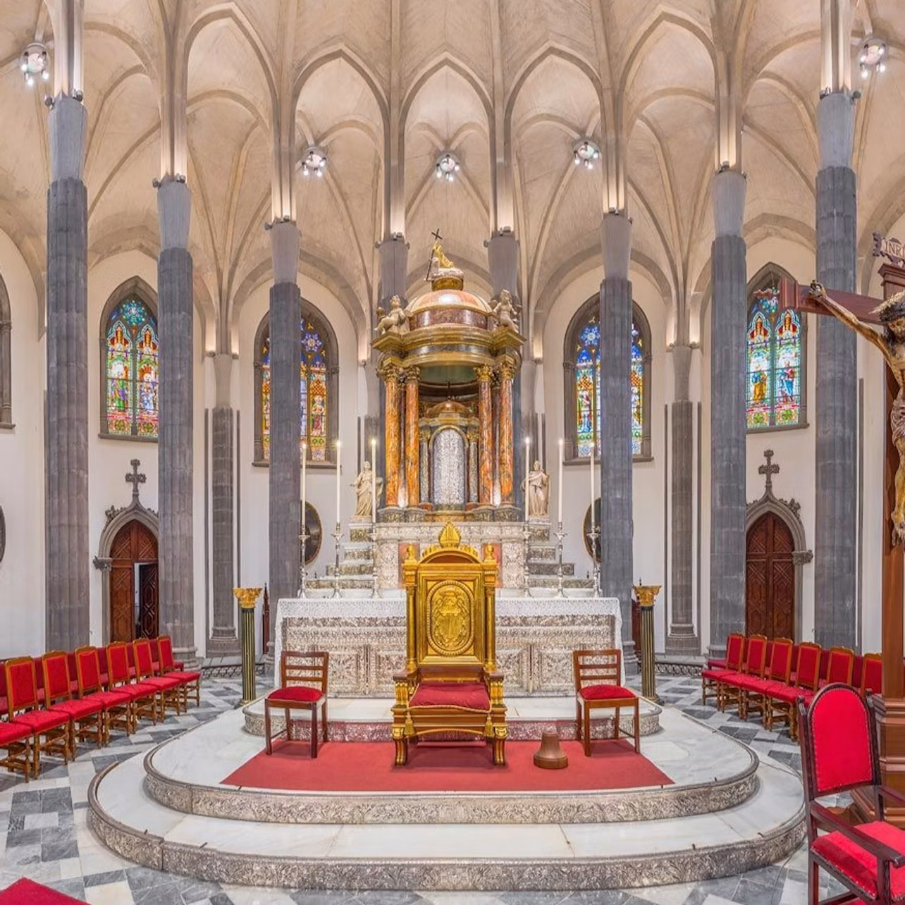 An ornate church interior with a central altar, stained glass windows, and red-seated chairs arranged in rows.