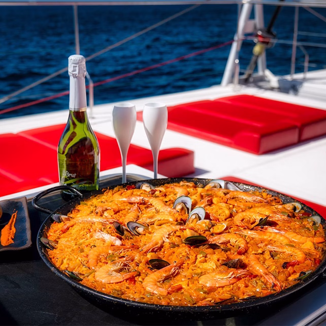 A seafood paella, champagne bottle, and two glasses on a yacht with red seating on a calm sea.