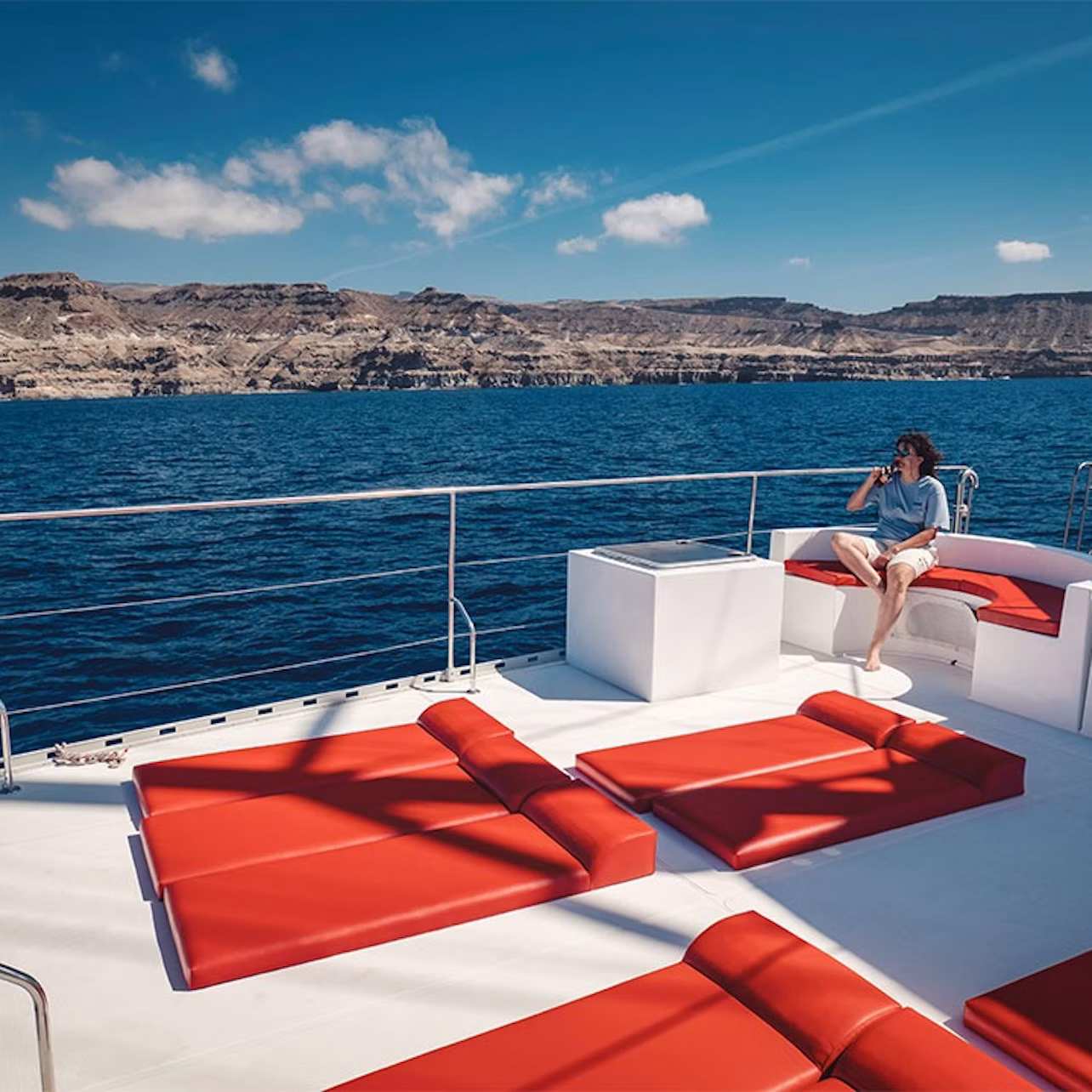 Person sitting on a boat deck with red cushions, overlooking calm blue water and rocky cliffs under a clear sky.