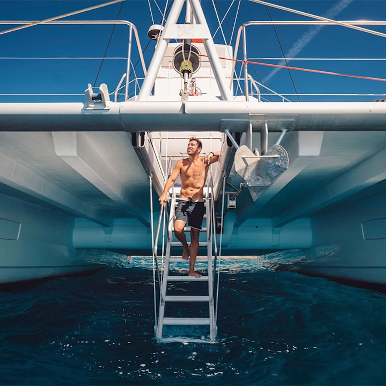 A man in swim shorts climbs a ladder on a large catamaran over clear blue water under a bright, sunny sky.