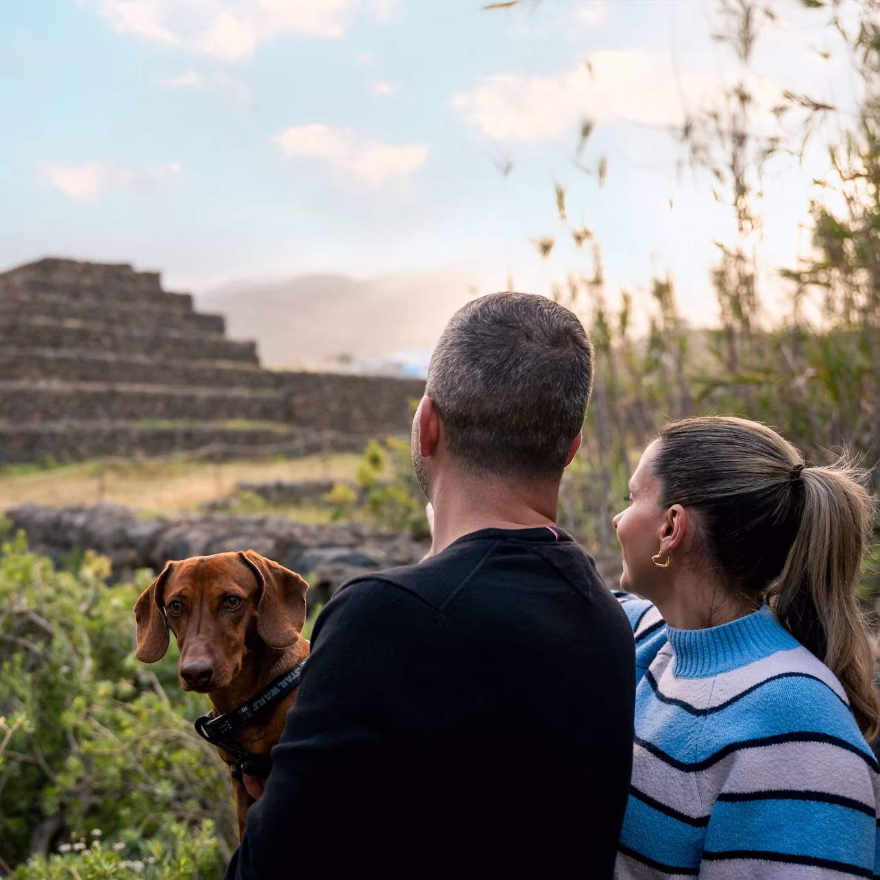 A man, a woman, and a dog look at a stepped stone structure in an outdoor setting with a cloudy sky.