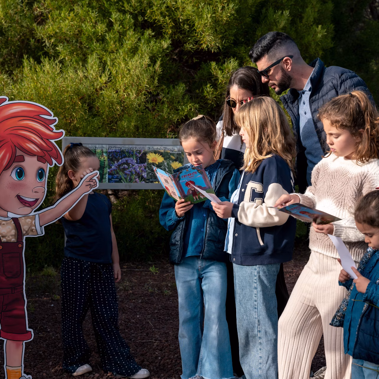 A group of children and an adult stand outdoors reading books near a colorful sign. Trees are visible in the background.