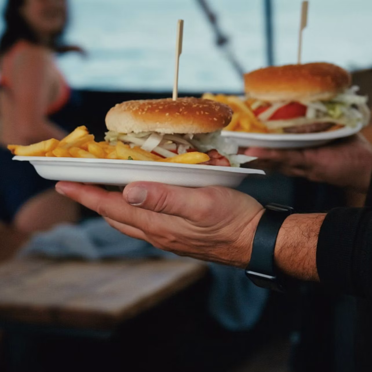 A person holding two plates with burgers and fries, with a blurred background of another person seated and a table.