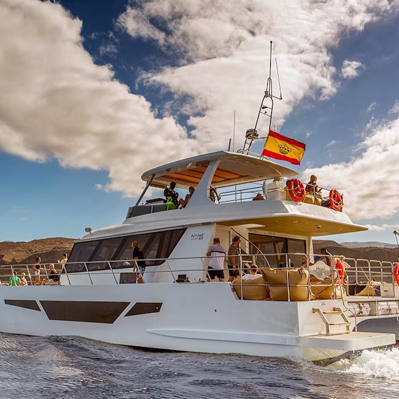 A large white yacht with people onboard, sailing on water. The yacht displays a red and yellow flag, under a partly cloudy sky.