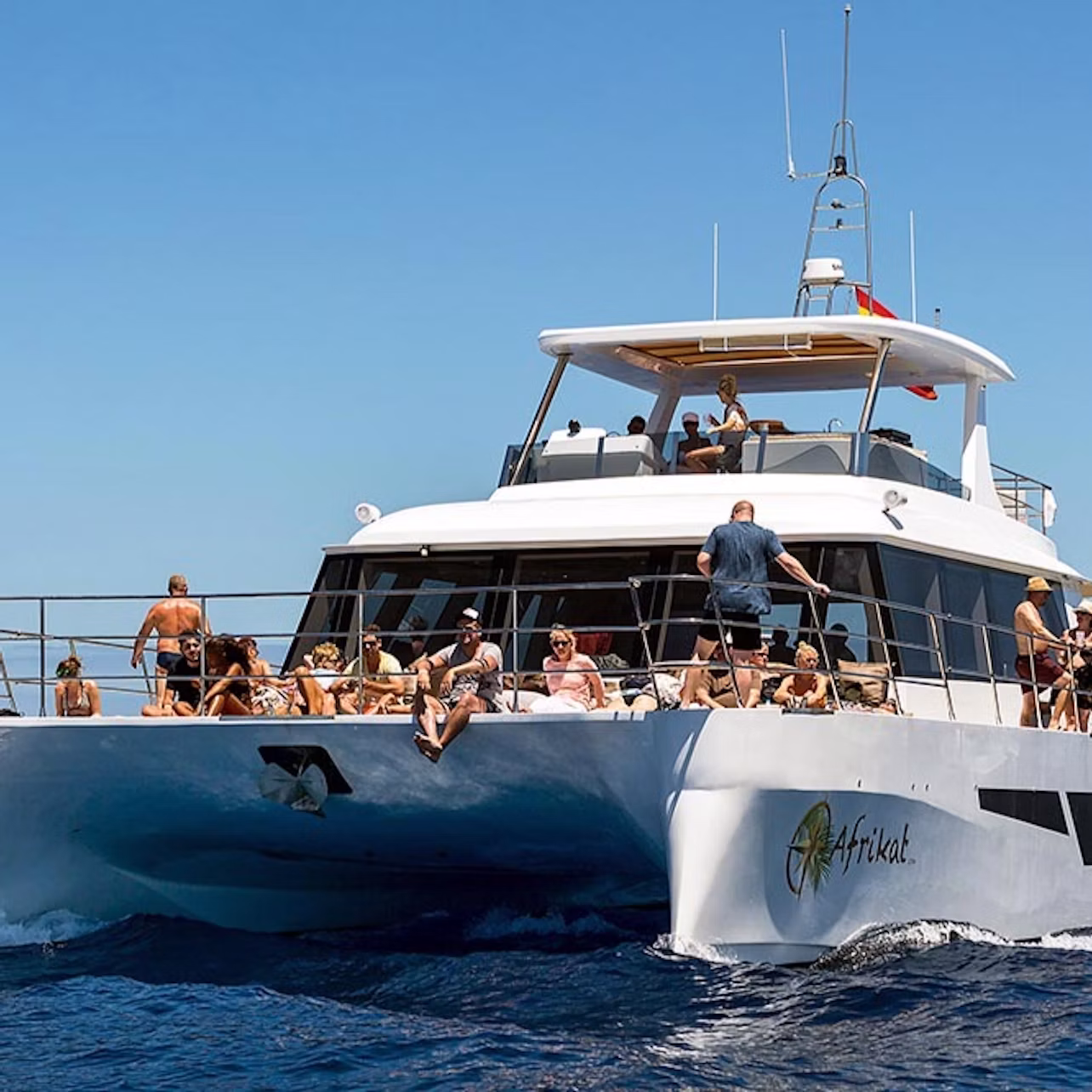 A group of people relaxing and sunbathing on the deck of a white yacht named "Afrikat," sailing on the blue ocean under a clear sky.