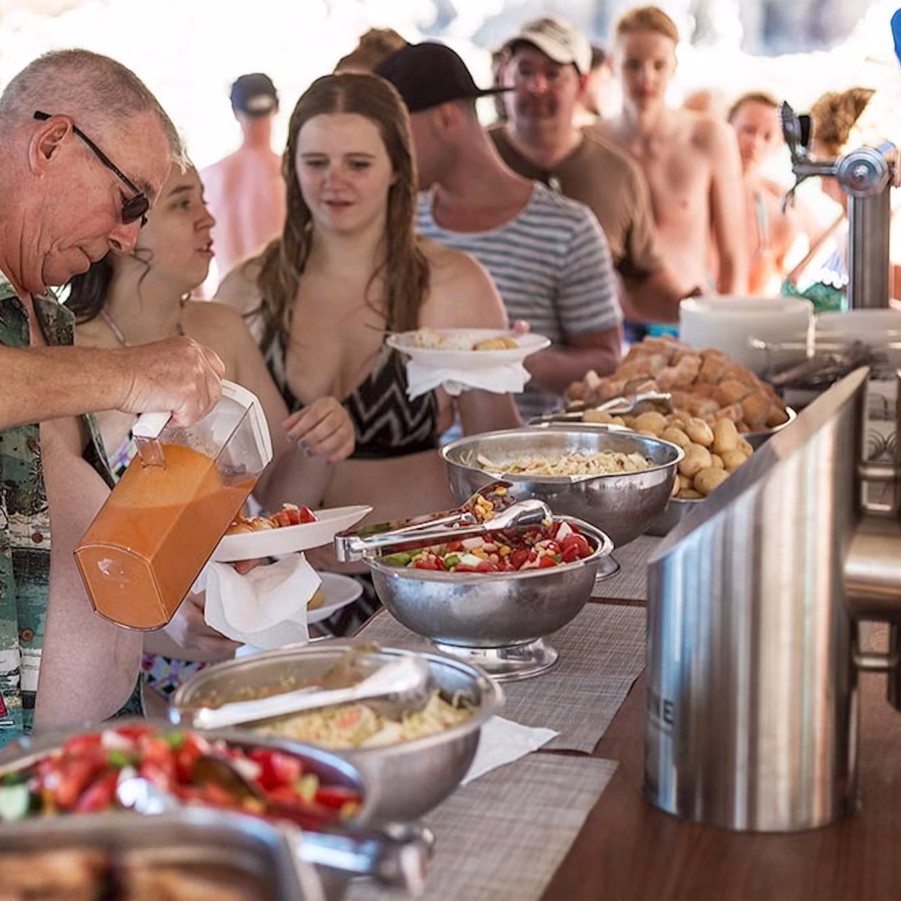 A group of people in swimwear are serving themselves food from a buffet with various dishes and a drink dispenser.