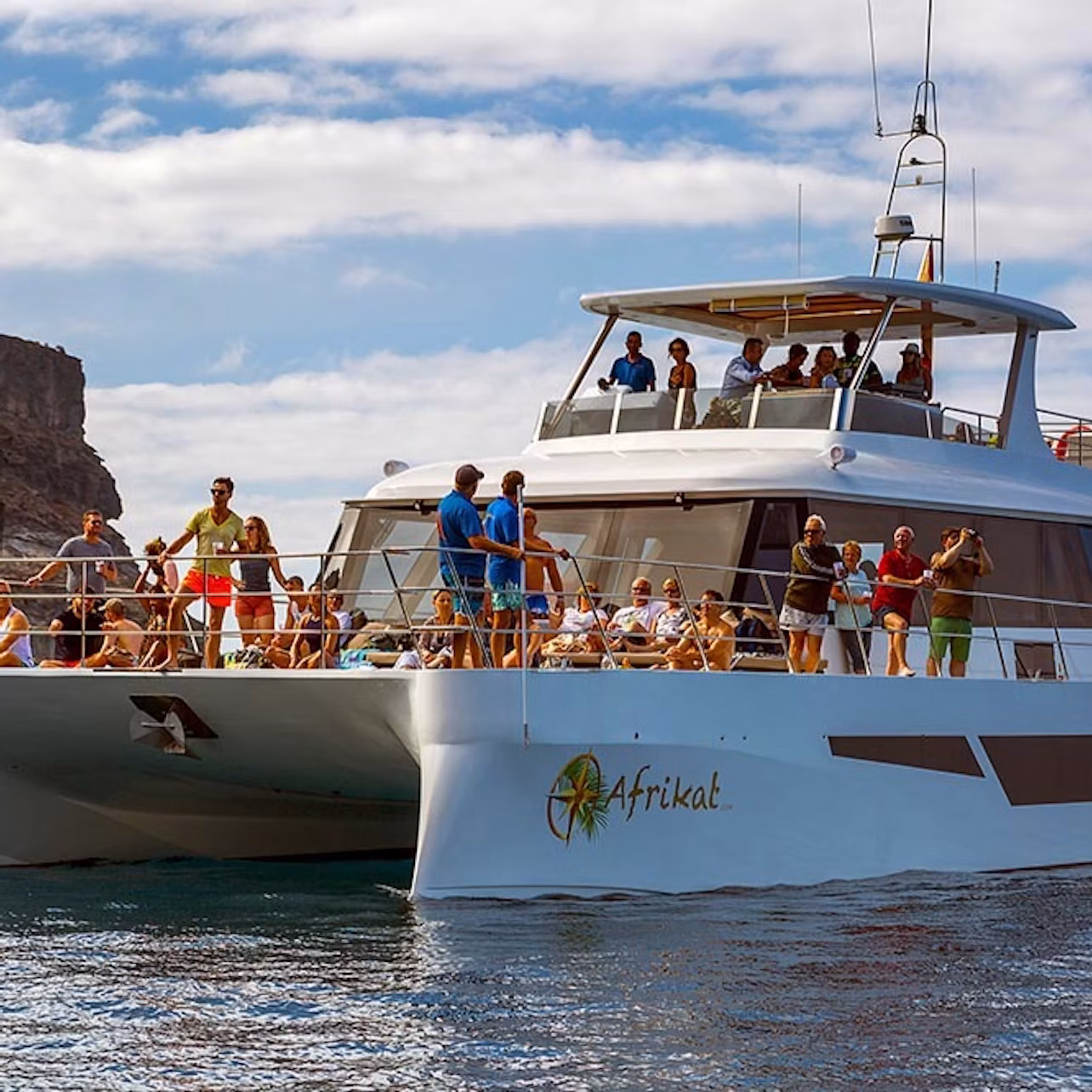 A large catamaran with people on board enjoying the sunny day on the water, with rocky cliffs and a partly cloudy sky in the background.