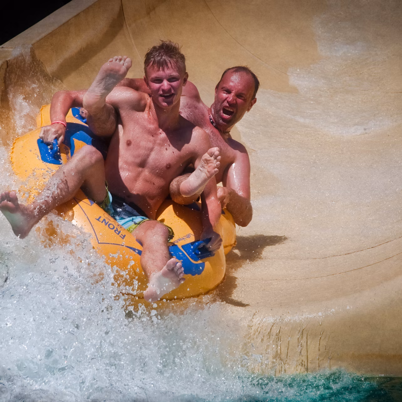 Two men on a yellow inflatable raft sliding down a water slide, with water splashing around them.