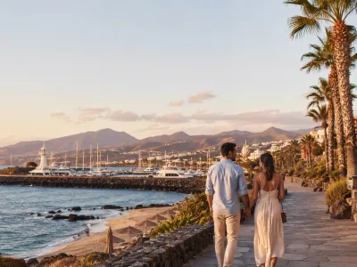 Couple walking along a Canary Islands seafront promenade at sunset with marina and beach views